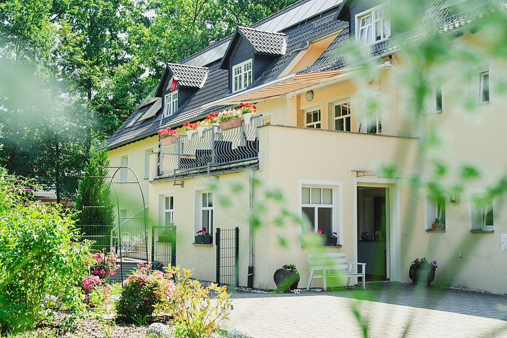 Cremefarbenes Haus mit Balkon voller roter Geranien, davor eine weiße Bank neben der offenen Haustür und ein blühender Garten