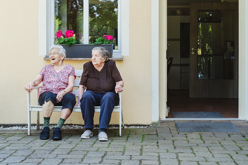Zwei Bewohnerinnen sitzen auf einer Bank neben der offenen Haustür, über ihnen ein Fensterkasten mit Geranien
