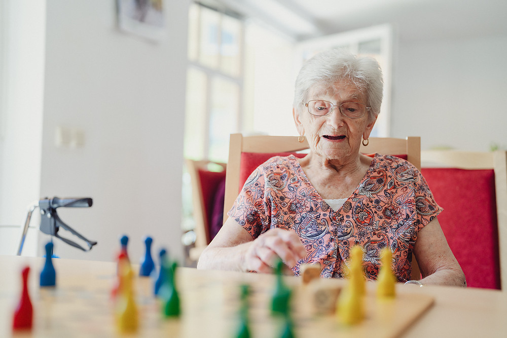 Bewohnerin sitzt am Tisch des Gemeinschaftsraums und spielt ein Brettspiel mit bunten Figuren