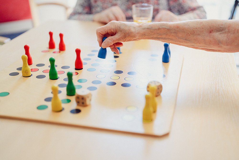 Ältere Hand setzt eine blaue Spielfigur auf ein Brettspiel mit bunten Kegeln