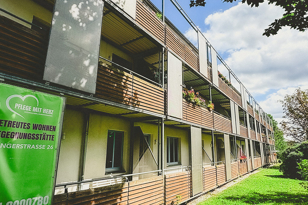 Wohnhaus Klingerstraße 26 mit Holzbalkonen voller Blumenkästen und dem Schild von Pflege mit Herz