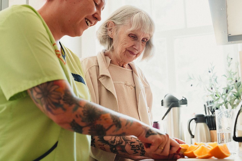 Eine Pflegekraft und eine ältere Dame schneiden gemeinsam lachend Obst in einer hellen Küche