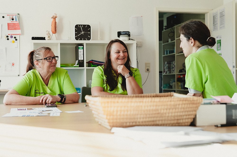 Drei Kolleginnen in grünen Poloshirts sitzen lachend an einem Tisch im hellen Büro und besprechen sich