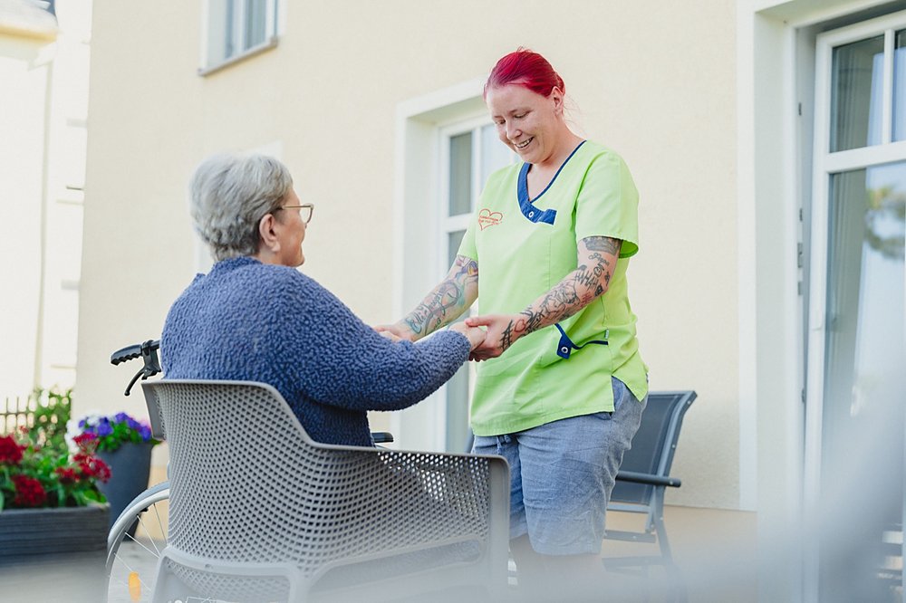 Eine Pflegekraft im grünen Kasack hält lächelnd die Hände einer Bewohnerin auf der Terrasse
