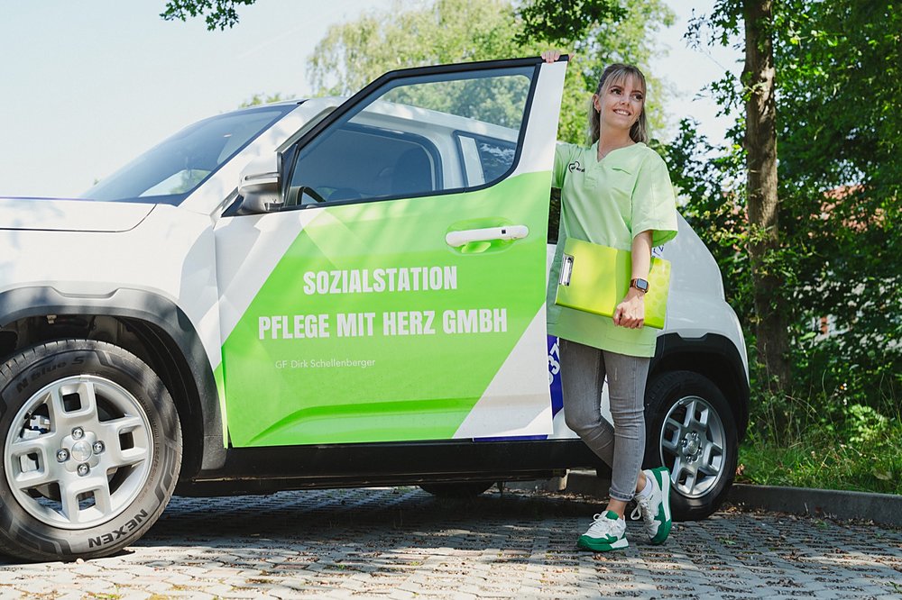 Pflegekraft in grüner Dienstkleidung steht mit ihrer Tourenmappe an der geöffneten Tür des beschrifteten Dienstwagens der Sozialstation Pflege mit Herz